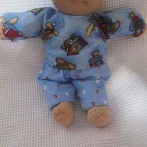 May include: A blue and white pajamas set with a bear and house print for a baby doll. The pajamas have a long-sleeved shirt and pants with a heart pattern.