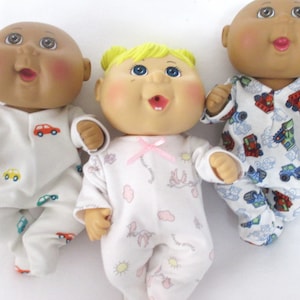 May include: Three Cabbage Patch Kids dolls wearing pajamas. The dolls are all different ethnicities and have different colored hair. The doll on the left is wearing a white and blue pajama with a car print. The doll in the middle is wearing a white and pink pajama with a cloud and bird print. The doll on the right is wearing a white and blue pajama with a truck print.