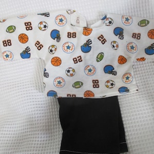 May include: A white baby outfit with a sports-themed pattern. The top features footballs, basketballs, soccer balls, and helmets, along with the numbers 88, 26, and 92. The bottom is solid black. The outfit is likely for a baby or toddler.