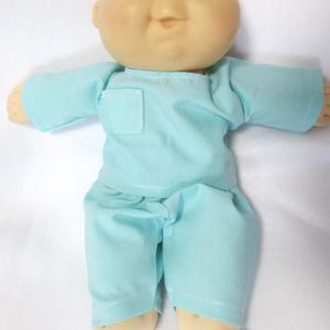 May include: A Cabbage Patch Kid doll with a round, pale face, blue eyes, and a small smile. The doll is wearing a light blue outfit, including a top with a pocket and matching pants. The doll's hands and feet are a light tan color.