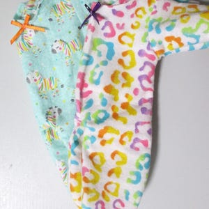 May include: Two baby onesies. One is light blue with colorful zebra print and an orange bow. The other is white with a rainbow leopard print and a purple bow. Both are made of soft fabric.