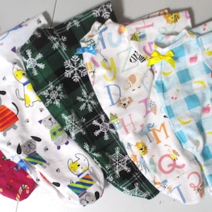 May include: A collection of baby onesies in various patterns. Designs include candy canes, dogs in sweaters, snowflakes, letters, and holiday scenes. Each onesie has a different color scheme, with bows at the neckline.