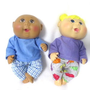 May include: Two Cabbage Patch Kids dolls wearing pajamas. One doll has brown skin and is wearing a blue shirt and blue and white plaid pants. The other doll has blonde hair and is wearing a purple shirt and gray pants with a colorful fruit print.