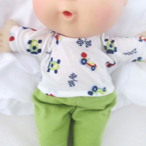 May include: A Cabbage Patch Kid doll wearing a white shirt with a blue, red, and green race car pattern and green pants. The doll has blonde hair and blue eyes.