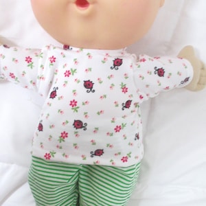 May include: A Cabbage Patch Kid doll wearing a white and green outfit with a ladybug print. The doll has blonde hair and blue eyes.