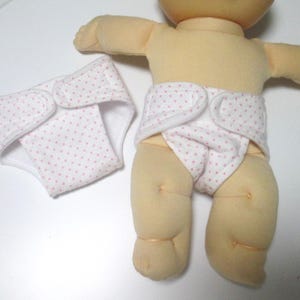 May include: A white doll diaper with pink polka dots. The diaper has a velcro closure and is made of soft fabric.