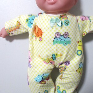 May include: A baby doll wearing a yellow and white checkered onesie with colorful cartoon animal designs. The onesie has a small light blue bow at the neckline. The doll has a smiling expression.
