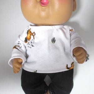 May include: A doll with a round head, brown eyes, and pink cheeks wearing a white long-sleeved shirt featuring a brown horse and green cactus print pattern, paired with solid black pants that have visible white stitching along the bottom hems.