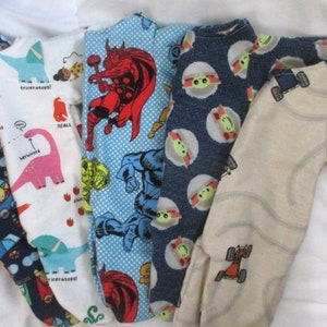 May include: Five different baby onesies with various patterns. One onesie is white with pink and green dinosaurs, another is blue with a green and blue superhero, another is blue with a green alien, another is beige with orange trucks, and the last one is navy blue with a yellow and red circus theme.