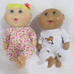 May include: Two Cabbage Patch Kids dolls wearing pajamas. One doll has blonde hair and is wearing pink and green pajamas with hearts. The other doll has brown skin and is wearing white pajamas with a brown horse design.