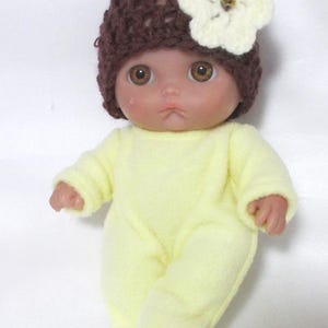 May include: A doll wearing a brown crocheted hat with a cream-colored flower and a yellow onesie. The doll has brown eyes and a neutral expression. The outfit is soft and cozy.