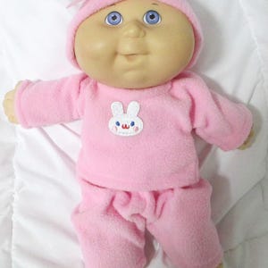 May include: A vintage Cabbage Patch Kids doll wearing a pink fleece outfit and matching hat with a bow. The doll has blue eyes, a smiling expression, and a white bunny applique on the chest.