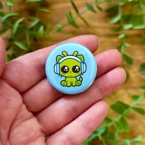 May include: A round, light blue button featuring a cartoon alien cat wearing headphones. The alien cat is green with large black eyes and yellow feet. The button is held in a hand, with green foliage in the background.