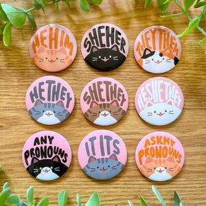 May include: Nine pink and white round buttons with different cat illustrations and text. The text on the buttons includes "He/Him", "She/Her", "They/Them", "He/They", "She/They", "He/She/They", "Any Pronouns", "It/Its", and "Ask My Pronouns".