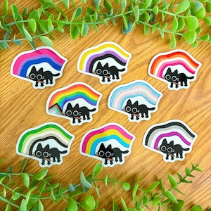 May include: Nine black cat stickers with different rainbow pride flags. Each sticker features a black cat holding a rainbow flag in its mouth.
