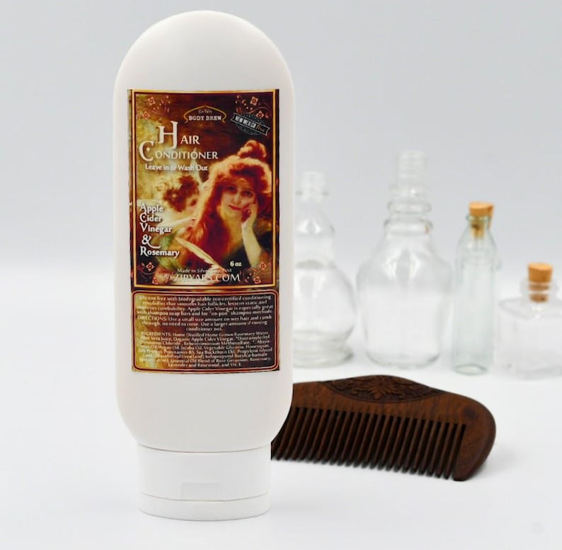 May include: A white bottle of hair conditioner with a vintage label featuring a woman with red hair. The label reads "Hair Conditioner Leave in or Wash Out Apple Cider Vinegar & Rosemary" and "SBODY BARN". The bottle is 6 oz and is made in the USA. The bottle is sitting on a white surface with a wooden comb.