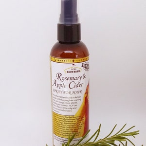 May include: A brown glass bottle with a spray nozzle, labelled "Zir Yale BODY BREW Rosemary & Apple Cider SPRAY FOR HAIR". The bottle is sitting on a white surface with a sprig of rosemary in the foreground.