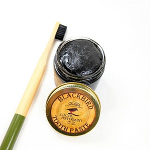 Peut inclure: Un pot de dentifrice Blackbird Cocoa Peppermint noir et or avec une brosse &agrave; dents noire en arri&egrave;re-plan.