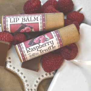 May include: Two lip balm tubes labelled "Lip Balm" and "Raspberry Truffle" sit on a decorative plate with fresh raspberries and chocolate pieces. The tubes have a brown paper casing with a pink and white label.