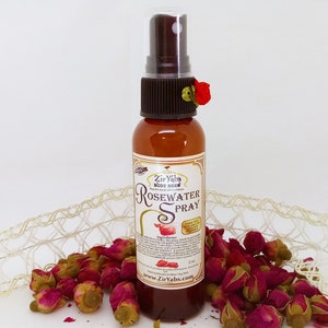 May include: A small brown bottle with a clear spray nozzle and a red flower on the top. The bottle is labeled "Zir Yabs Body Brew Rosewater Spray" and has a list of ingredients on the label. The bottle is sitting on a white surface with dried rose buds.