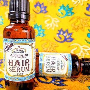 Pu&ograve; includere: Due bottiglie di vetro ambrato con contagocce, etichettate "Andalusian Precious Oils Hair Serum" in un bordo blu e dorato. Le bottiglie sono appoggiate su un tessuto giallo con un motivo floreale.
