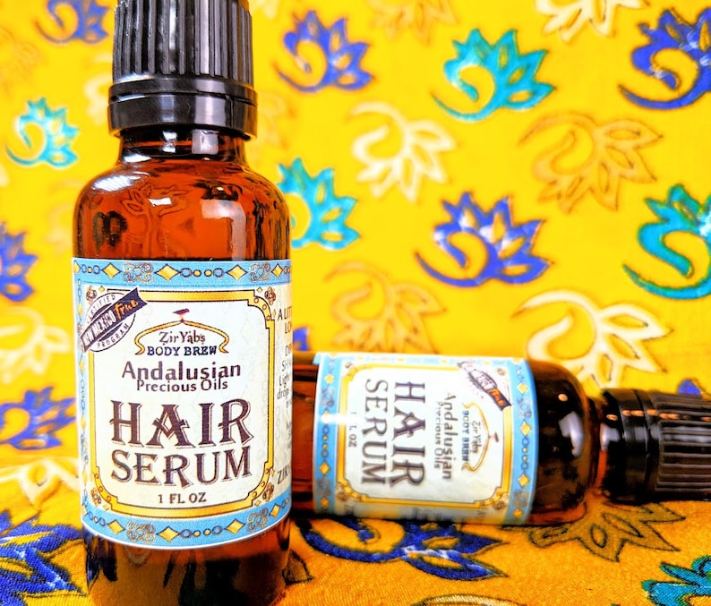 Pu&ograve; includere: Due bottiglie di vetro ambrato con contagocce, etichettate "Andalusian Precious Oils Hair Serum" in un bordo blu e dorato. Le bottiglie sono appoggiate su un tessuto giallo con un motivo floreale.
