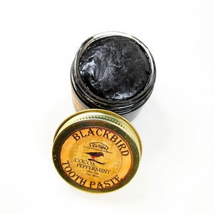 Peut inclure: Un pot dor&eacute; avec un couvercle noir contenant du dentifrice noir. Le couvercle porte une &eacute;tiquette qui dit "Blackbird" avec une petite illustration d'oiseau et le texte "Cocoa Peppermint Tooth Paste".
