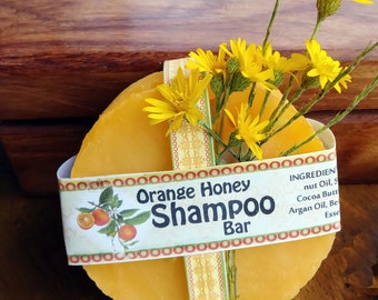May include: A round bar of orange honey shampoo wrapped in a yellow and orange patterned paper with a label that reads "Orange Honey Shampoo Bar". The label also lists the ingredients: nut oil, cocoa butter, argan oil, and essential oils. The bar is decorated with a sprig of yellow flowers.