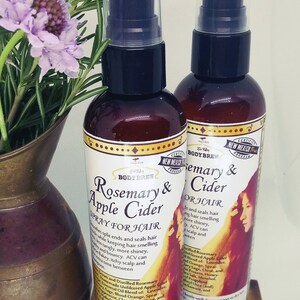 May include: Two brown glass bottles with clear pump tops, labelled "Rosemary & Apple Cider Spray for Hair" and "Rosemary & Cider for Hair". The bottles are sitting on a wooden surface with a copper vase and rosemary sprigs in the background.