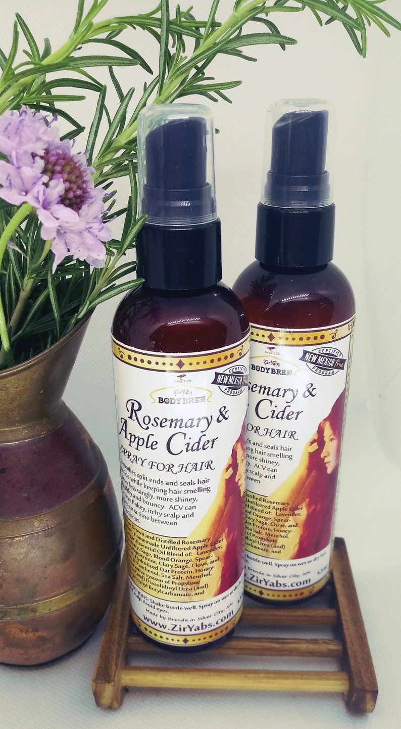 May include: Two brown glass bottles with clear pump tops, labelled "Rosemary & Apple Cider Spray for Hair" and "Rosemary & Cider for Hair". The bottles are sitting on a wooden surface with a copper vase and rosemary sprigs in the background.