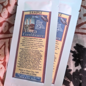 May include: A white sample packet of Yucca Conditioner hair care product with a brown and red border. The packet features a green plant illustration and the text "SAMPLE", "Zir Yabs", "Yucca Conditioner", "Desert inspired hair care with protective yucca juice!" and "Made in Silver City, NM ZIRYABS.COM The yucca flower is the state flower of New Mexico."