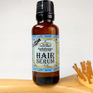 Pu&ograve; includere: Una bottiglia di vetro marrone con un tappo nero, etichettata "Zir Yabs Body Brew Andalusian Precious Oils Hair Serum 1 FL OZ".