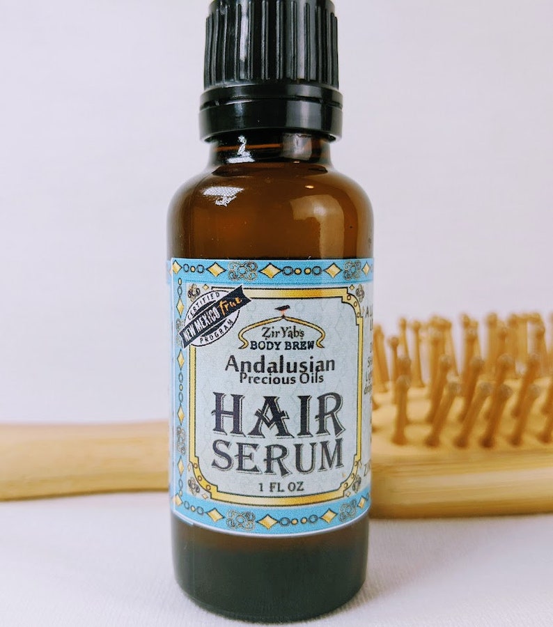 Pu&ograve; includere: Una bottiglia di vetro marrone con un tappo nero, etichettata "Zir Yabs Body Brew Andalusian Precious Oils Hair Serum 1 FL OZ". L'etichetta &egrave; blu e gialla con un bordo decorativo. La bottiglia &egrave; su una superficie bianca.