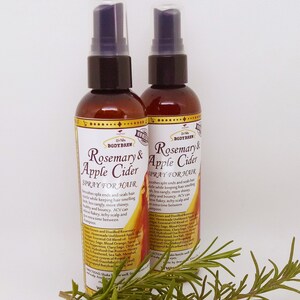 May include: Two brown glass bottles with gold labels and black spray tops. The labels read "Rosemary & Apple Cider Spray for Hair". The bottles are sitting on a green rosemary sprig.