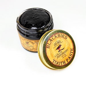 Peut inclure: Un pot en verre de dentifrice au charbon de bois noir avec un couvercle dor&eacute;. Le couvercle porte le texte "Blackbird Zir Vabs for easy & natural teeth care" et "Cocoa Peppermint Tooth Paste".
