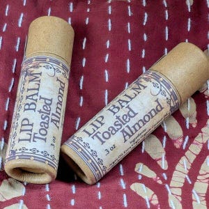 May include: Two lip balm tubes with a Toasted Almond flavor. The tubes are cylindrical with a brown cap and a label that reads "Lip Balm Toasted Almond .3 oz". The background is a burgundy fabric.