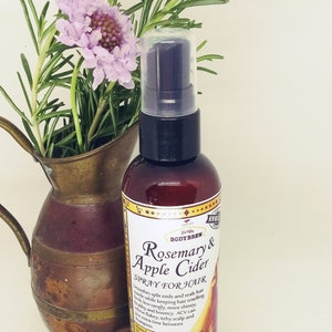 May include: A brown glass bottle with a gold label that reads "Rosemary & Apple Cider Spray for Hair". The bottle is sitting on a copper-coloured pitcher with a sprig of rosemary and a purple flower.