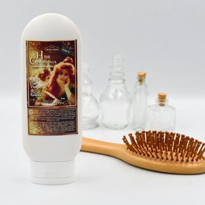 May include: White plastic bottle of hair conditioner with a label that says "Hair Conditioner" and "Apple Cider Vinegar & Rosemary". The bottle is sitting on a white surface with a wooden hairbrush.