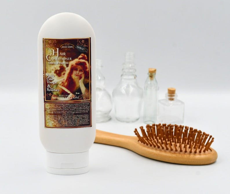 May include: White plastic bottle of hair conditioner with a label that says "Hair Conditioner" and "Apple Cider Vinegar & Rosemary". The bottle is sitting on a white surface with a wooden hairbrush.