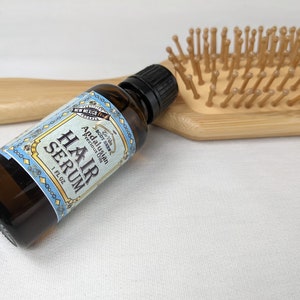 Pu&ograve; includere: Un flacone di siero per capelli in vetro marrone con un'etichetta blu su cui &egrave; scritto "Hair Serum" e "Andalusian Precious Oil". Il flacone &egrave; appoggiato su una superficie bianca accanto a una spazzola per capelli in legno.