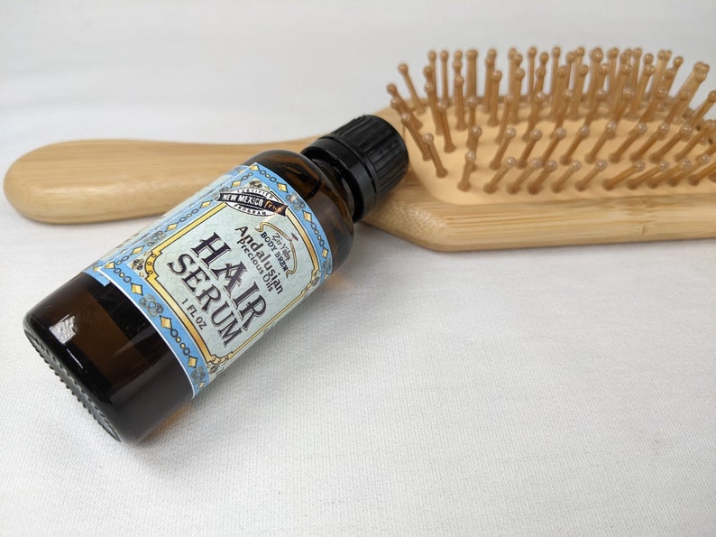 Pu&ograve; includere: Un flacone di siero per capelli in vetro marrone con un'etichetta blu su cui &egrave; scritto "Hair Serum" e "Andalusian Precious Oil". Il flacone &egrave; appoggiato su una superficie bianca accanto a una spazzola per capelli in legno.