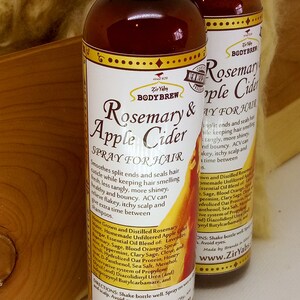 May include: Two amber glass bottles with gold labels and a red apple design. The labels read "Rosemary & Apple Cider Spray for Hair" and list ingredients including rosemary, apple cider vinegar, and essential oils. The bottles are on a wooden surface.