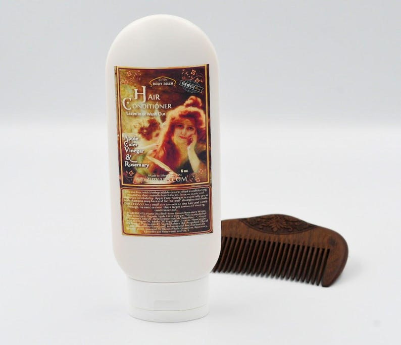 May include: White bottle of hair conditioner with a vintage label featuring a woman with red hair. The label reads "Hair Conditioner Leave-in Treatment" and "Apple Cider Vinegar & Rosemary". The bottle is next to a wooden comb.