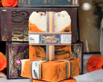 May include: Three bars of handmade soap stacked on top of each other. The top bar is white with orange swirls and has a label that says "Pumpkin". The middle bar is brown and orange with swirls and the bottom bar is orange. The soap is sitting on a table with a book and a bottle in the background.