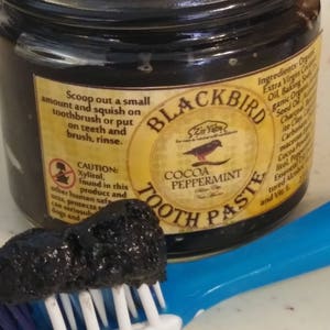 Peut inclure: Un pot en verre de dentifrice &agrave; la menthe poivr&eacute;e et au cacao Blackbird Zir Yabs. Le pot est jaune et noir avec un logo d'oiseau noir. Le dentifrice est de couleur charbon fonc&eacute;. Une brosse &agrave; dents bleue avec une t&ecirc;te de poils blancs a une noisette de dentifrice dessus.