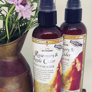 May include: Two brown glass bottles with spray tops, labelled "Rosemary & Apple Cider Spray for Hair" and "Rosemary & Cider Spray for Hair". The bottles are sitting on a wooden surface with a copper vase and purple flowers.