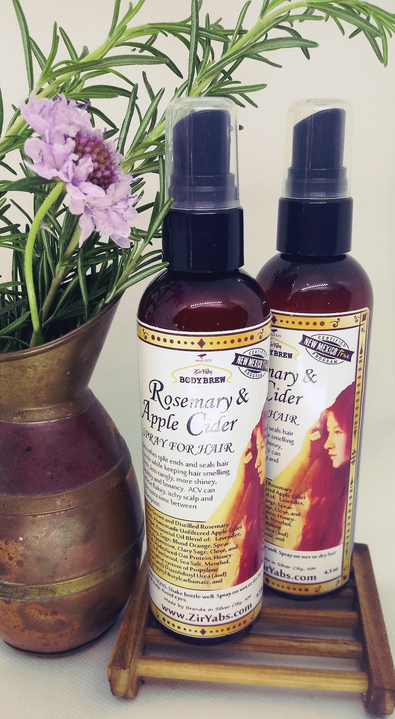 May include: Two brown glass bottles with spray tops, labelled "Rosemary & Apple Cider Spray for Hair" and "Rosemary & Cider Spray for Hair". The bottles are sitting on a wooden surface with a copper vase and purple flowers.