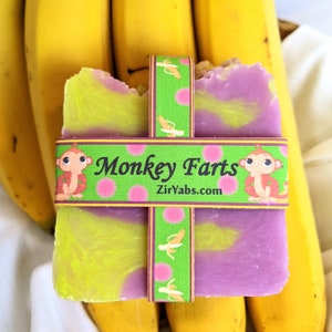 May include: A purple and yellow bar of soap with a green and pink label that says "Monkey Farts" and "ZirYabs.com". The soap is sitting on top of a bunch of yellow bananas.