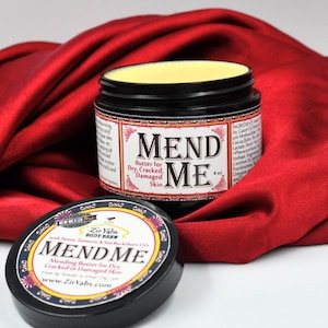 以下が含まれることがあります： 乾燥、ひび割れ、損傷した肌のための「Mend Me」ボディバター4オンス入り。ジャーは開いており、バターは薄い黄色です。ジャーは赤い布の背景の上にあります。