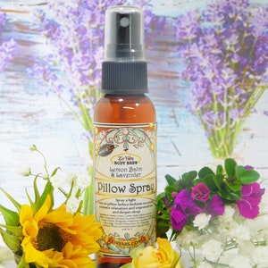 May include: A small brown bottle with a spray nozzle, labeled "Zir Yaby BODY BREW Lemon Balm & Lavender Pillow Spray". The bottle is surrounded by flowers, including lavender, sunflowers, and roses.